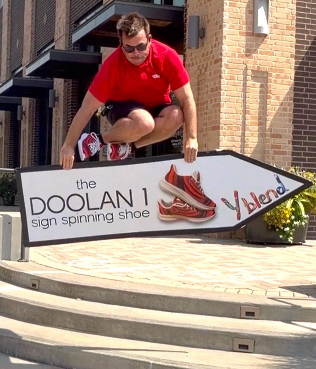 A man in a red t-shirt and sunglasses jumps over a large white directional sign that reads 'the DOOLAN 1 sign spinning shoe' with a red sneaker graphic and the word 'Y blend'. The scene is set outdoors on stone steps in front of a brick building.